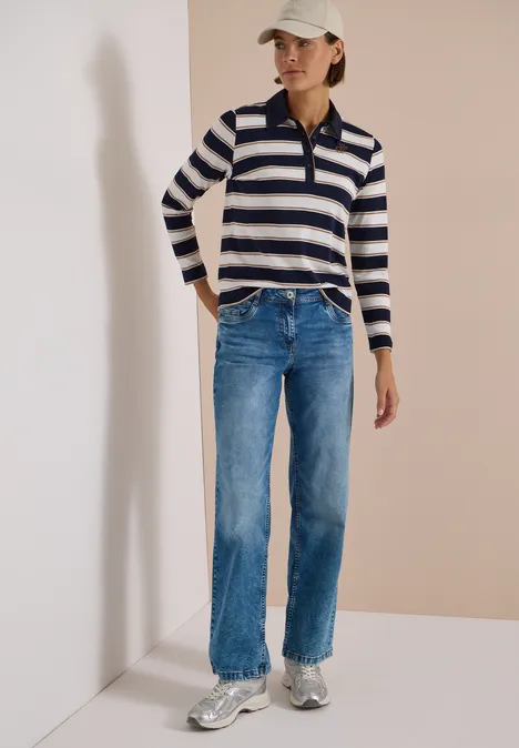 Femme en pull rayé, jean bleu, casquette blanche et baskets sur fond neutre.