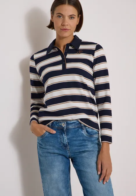 Femme portant un polo à manches longues rayé bleu et beige avec un jean, devant un fond neutre.