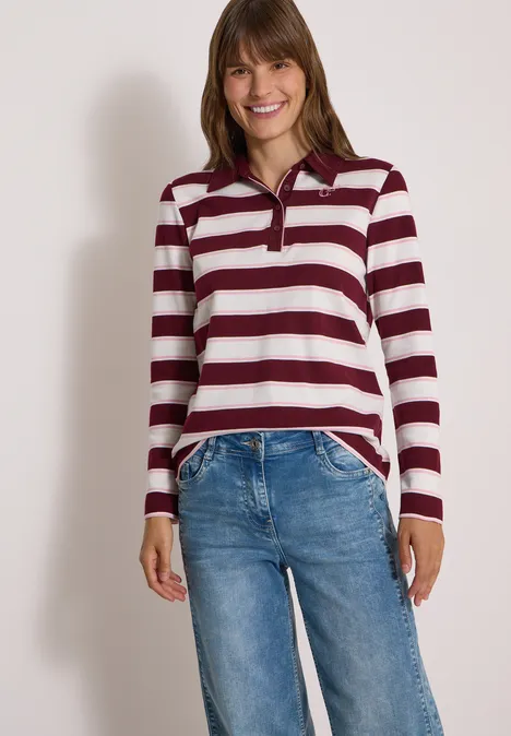 Junge Frau lächelnd in einem burgund-weiß gestreiften Langarmshirt und blauen Jeans.