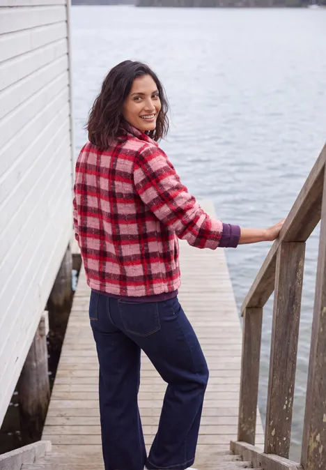 Eine Frau in kariertem roten Pullover und Jeans steht lächelnd auf einem Steg am See.