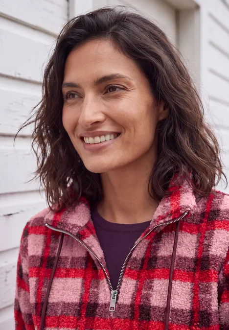 Eine lächelnde Frau in einem rot-karierten Fleecejacke vor einer weißen Hauswand.
