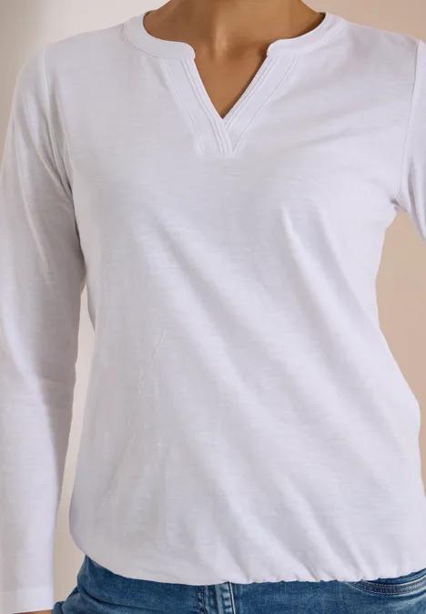 Oberkörper einer Frau in schlichtem weißen Langarmshirt mit V-Ausschnitt.