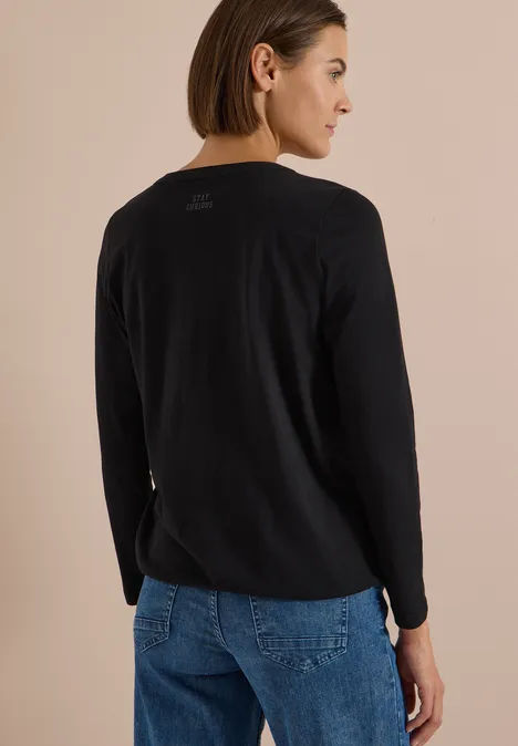 Eine Frau von hinten in schwarzem Pullover und blauen Jeans vor beigem Hintergrund.