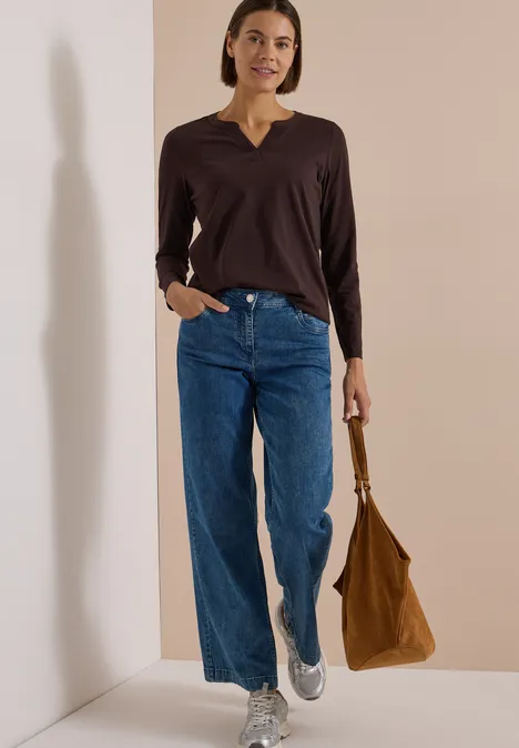 Frau in brauner Bluse, weiten Jeans und weißen Sneakern hält eine braune Tasche.