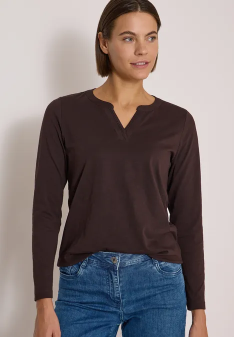 Frau in braunem Langarmshirt und blauer Jeans, kurzhaarig, stehend und zur Seite blickend.