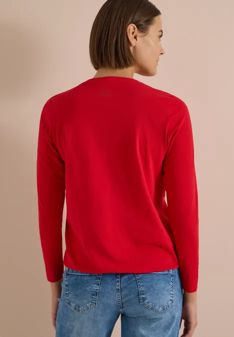 Frau von hinten in rotem Langarmshirt und blauer Jeans vor neutralem Hintergrund.