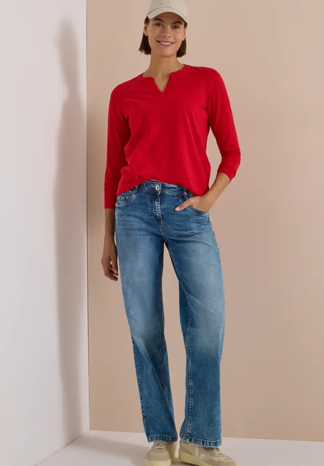 Frau in rotem Langarmshirt und blauen Jeans steht vor schlichtem Hintergrund und lächelt.