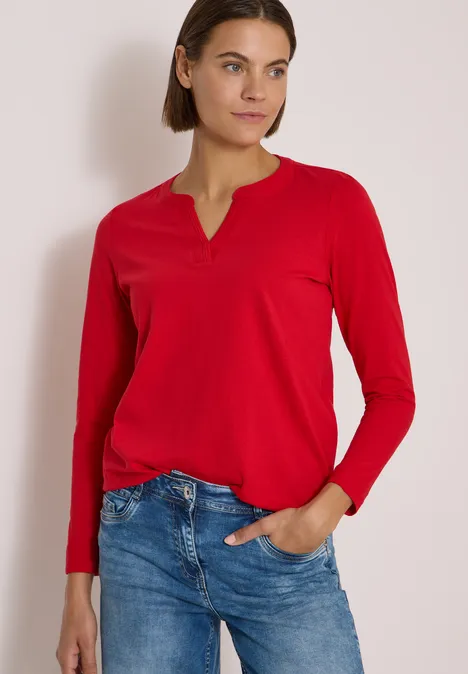 Eine Frau in rotem Langarmshirt und blauen Jeans steht vor hellem Hintergrund, eine Hand in der Hosentasche.