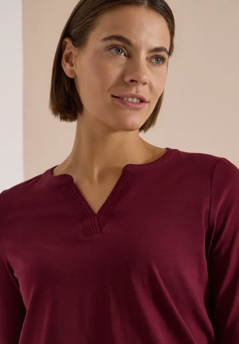 Eine Frau in einem weinroten Langarmshirt mit V-förmigem Ausschnitt vor hellem Hintergrund.