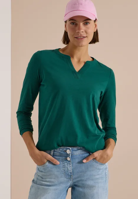 Frau mit grünem V-Shirt, blauen Jeans und rosa Cap steht lässig mit Händen in Hosentaschen.
