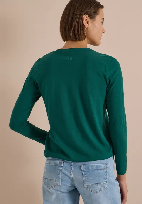 Eine Frau steht in grünem Langarmpullover und Jeans vor neutralem Hintergrund, Rückenansicht.