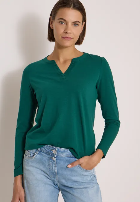 Eine Frau trägt ein grünes Langarmshirt und blaue Jeans, steht vor neutralem Hintergrund.
