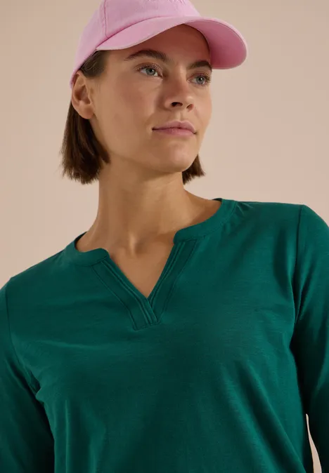 Eine Frau mit kurzem braunen Haar trägt ein grünes Shirt und eine rosa Baseballkappe, Blick seitlich.