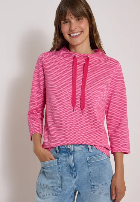 Frau trägt pinken, gemusterten Hoodie mit Dreiviertelärmeln und lockere blaue Jeans, lächelnd.