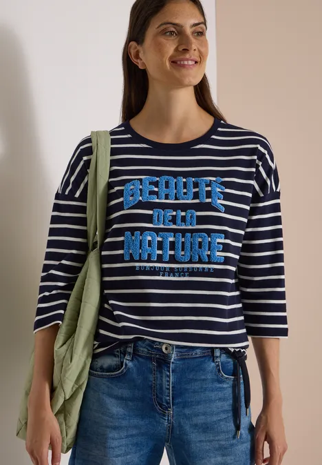 3/4-Arm Shirt Boat Neck und Streifen urban dark blue Frau in gestreiftem Shirt mit „Beauty of Nature“-Aufdruck, Jeans und grüner Jacke über der Schulter.
