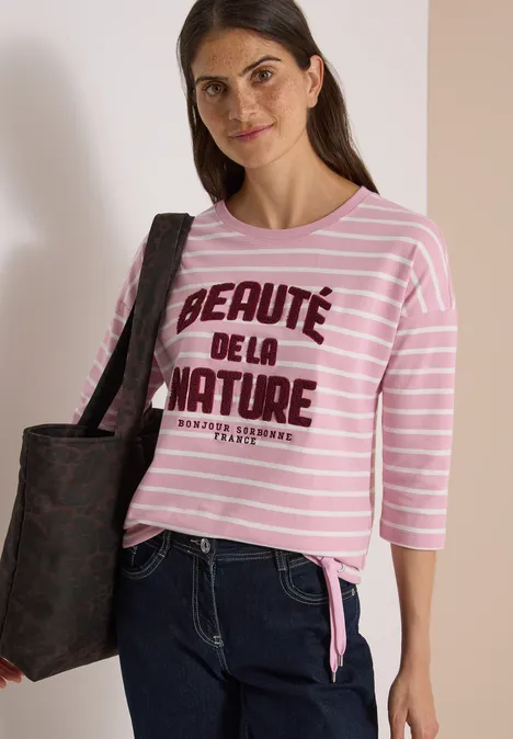 Frau trägt rosa-weiß gestreiftes Shirt mit Aufschrift 'Beauté de la nature', Jeans und schwarze Tasche.