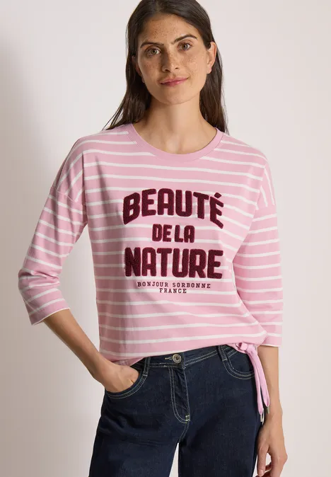 Eine lächelnde Frau trägt ein rosa-weiß gestreiftes Shirt mit Aufschrift 'Beauté de la nature' und Jeans.