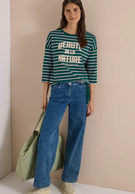 Frau mit gestreiftem Shirt, weiten Jeans und grünem Stoffbeutel vor beigem Hintergrund.