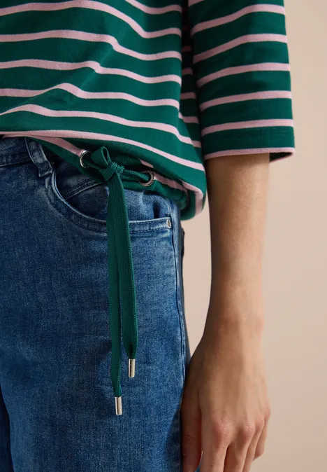 Person trägt grün-weiß gestreiftes Oberteil und blaue Jeans mit grünem Taillengürtel, Hand sichtbar