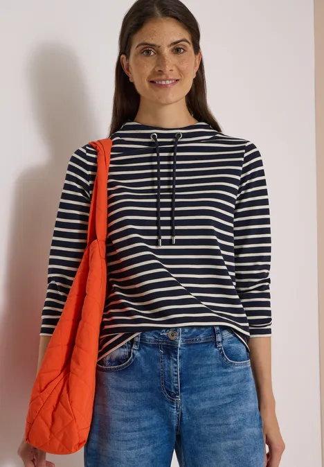 Langarm Shirt mit Funnel Neck und Streifen urban dark blue Frau mit gestreiftem Shirt, Jeans und orangefarbener Tasche lächelt vor hellem Hintergrund.