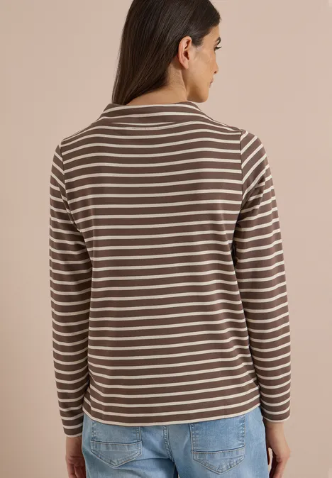 Femme de dos portant un haut à manches longues marron à rayures blanches et un jean bleu sur fond clair.