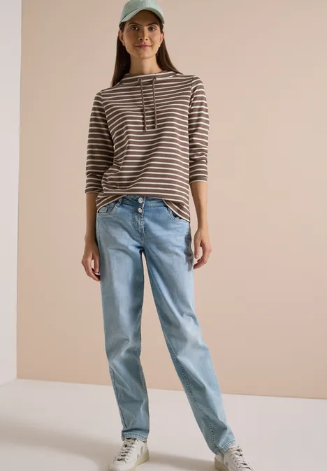 Femme portant un sweat à capuche rayé, un jean et une casquette sur fond beige.