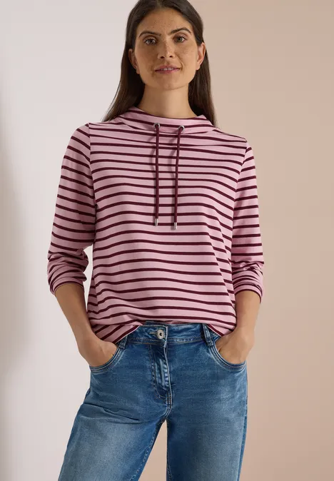 Frau in rosa-schwarz gestreiftem Kapuzenpullover und blauen Jeans vor neutralem Hintergrund.