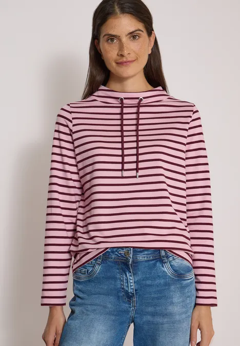 Junge Frau lächelt in pink-schwarz gestreiftem Hoodie und blauer Jeans vor neutralem Hintergrund.
