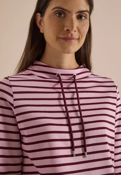 Frau in rosa-gestreiftem Hoodie mit Kordeln, dunklem Haar und schlichten Ohrringen.
