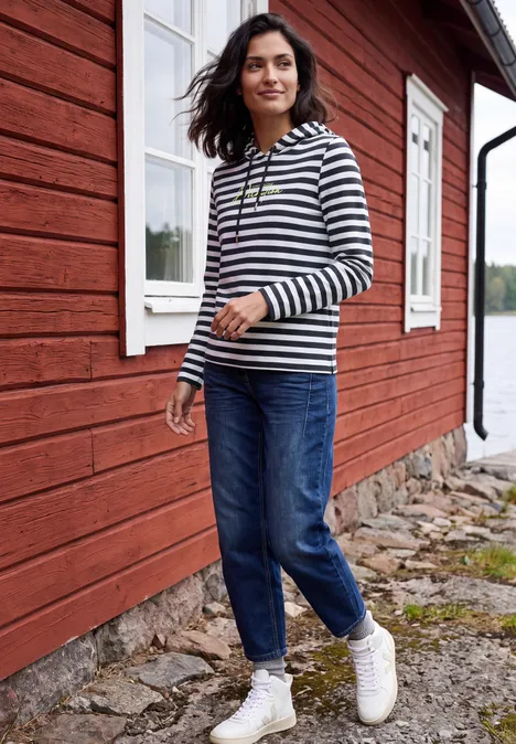 Langarm Shirt mit Kapuze und Streifen urban dark blue Frau in gestreiftem Pulli und Jeans spaziert vor einem roten Holzhaus am Wasser.