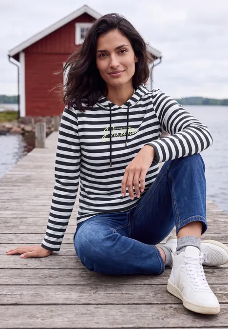 Langarm Shirt mit Kapuze und Streifen urban dark blue Frau in gestreiftem Hoodie und Jeans sitzt entspannt auf einem Steg am Wasser vor einem roten Bootshaus.