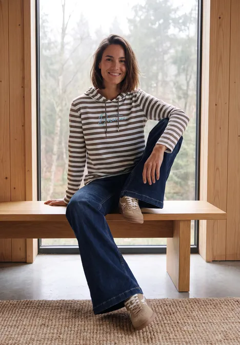 Vrouw zit ontspannen op een houten bank voor een raam, in een gestreepte trui en jeans.
