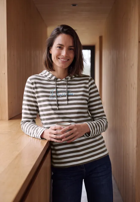Een glimlachende vrouw leunt ontspannen op een houten balustrade in een gang, met een gestreepte trui.