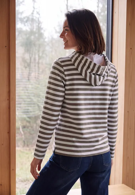 Vrouw in gestreepte capuchontrui staat bij het raam en kijkt naar het bos.
