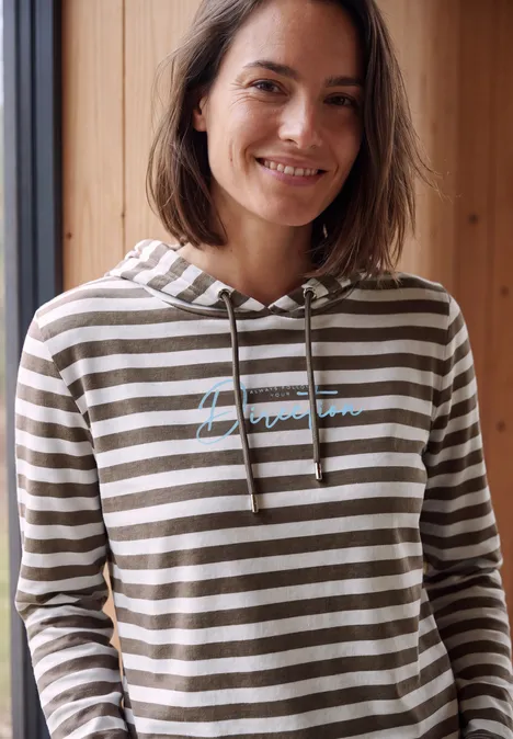 Een glimlachende vrouw in een gestreepte capuchonsweater voor een raam met houten panelen.
