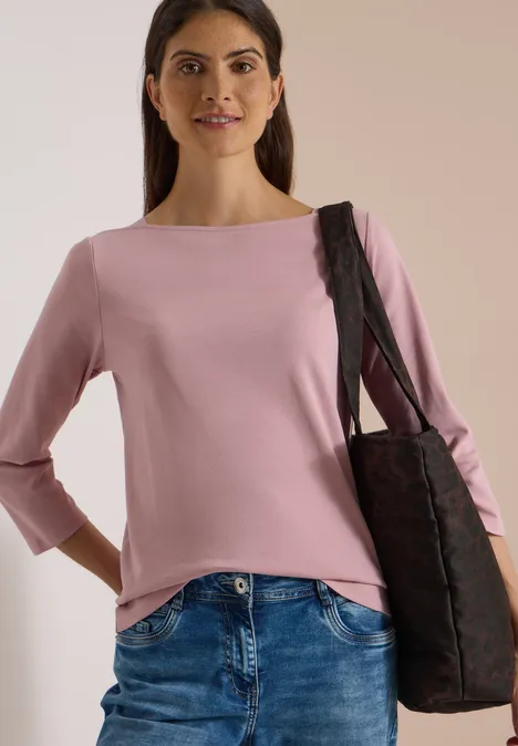 Frau in rosa Oberteil, Jeans und schwarzer Tasche vor neutralem Hintergrund.