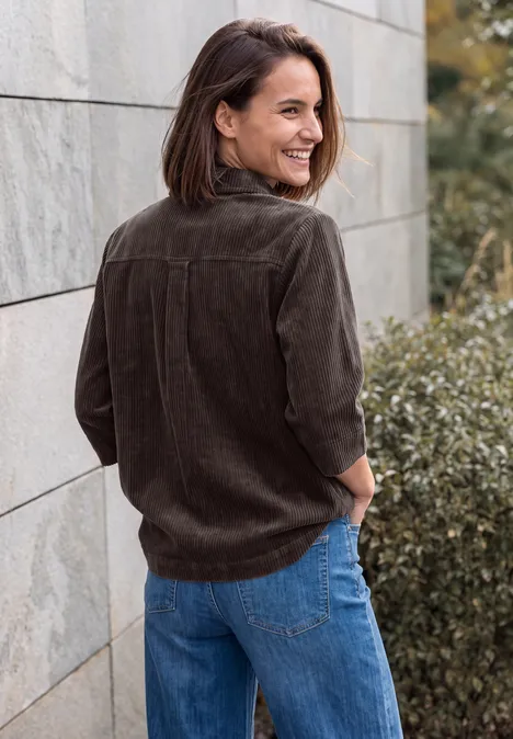 Jersey-corduroyblouse met halflange mouwen in effen kleur truffle brown Eine Frau in brauner Cordjacke und blauer Jeans lehnt lächelnd an einer Steinmauer im Freien.