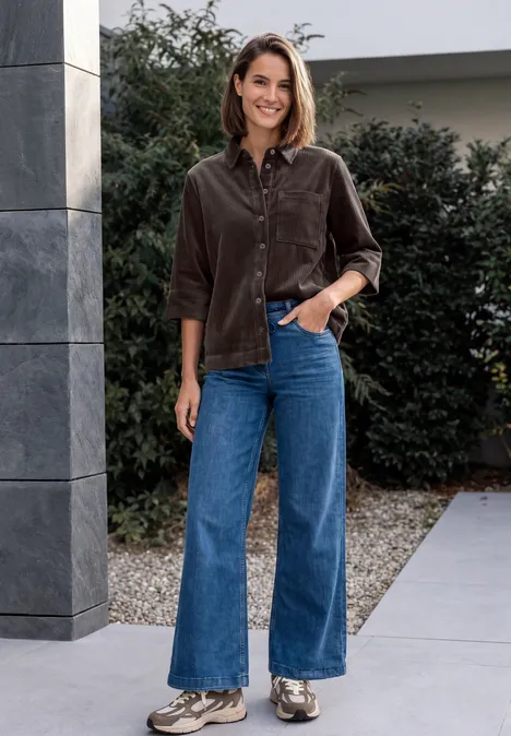 Jersey-corduroyblouse met halflange mouwen in effen kleur truffle brown Eine Frau trägt braunes Cord-Hemd und blaue Schlagjeans, steht lächelnd vor einem Gebäude im Freien.