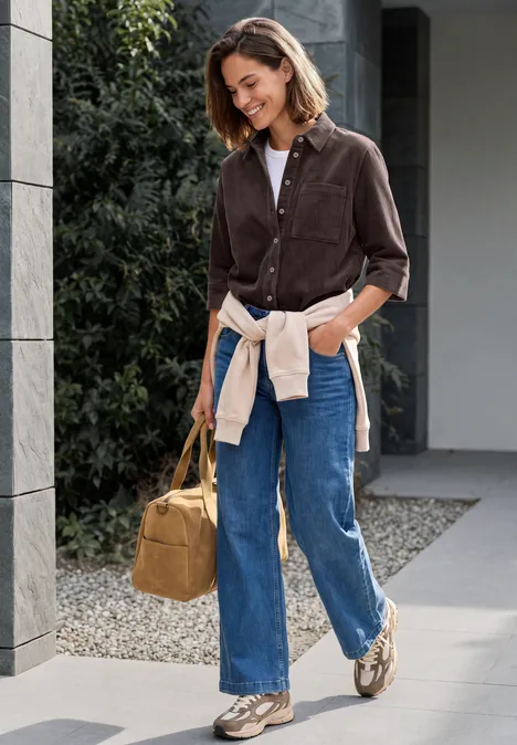 Jersey-corduroyblouse met halflange mouwen in effen kleur truffle brown Lässig gekleidete Frau mit Tasche spaziert lächelnd an einem modernen Gebäude entlang.