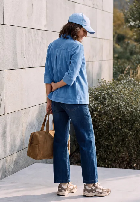 Jersey-corduroyblouse met halflange mouwen in effen kleur north blue Vrouw in lichtblauw jasje, spijkerbroek en pet staat zijwaarts tegen een muur en houdt een bruine tas achter haar rug.