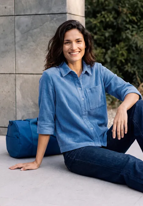 Jersey-corduroyblouse met halflange mouwen in effen kleur north blue Een glimlachende vrouw zit ontspannen voor een muur, draagt een denimoverhemd, met een blauwe tas naast zich.
