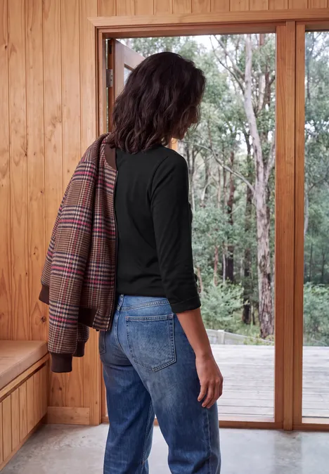 Shirt met lange mouwen en trekkoord aan de zoom Black Een vrouw in zwart topje en jeans draagt een jas over de schouder en kijkt door een open houten deur naar het bos.