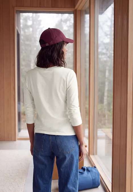 Langarm Shirt mit Tunnelzug am Saum vanilla white Eine Frau steht mit Jeans, weißem Shirt und roter Kappe vor einem Fenster und hält eine blaue Tasche.