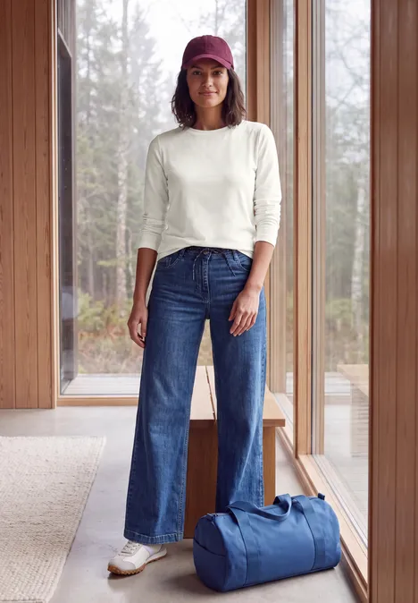 Langarm Shirt mit Tunnelzug am Saum vanilla white Frau mit roter Mütze, weißem Pullover und weiten Jeans steht neben blauer Tasche vor großen Fenstern im Holzhaus.