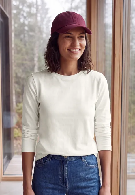 Langarm Shirt mit Tunnelzug am Saum vanilla white Frau in weißem Langarmshirt, Jeans und roter Mütze steht lächelnd vor großen Fenstern mit Waldblick.