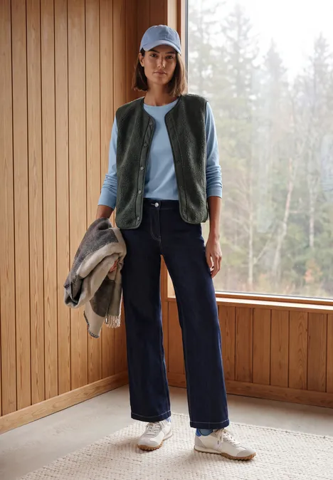 Langarm Shirt mit Tunnelzug am Saum cosmopolitan blue Frau im Casual-Look mit Weste, Cap, Jacke in der Hand, weiten Jeans und Sneakern in einem Holzraum vor Wald.