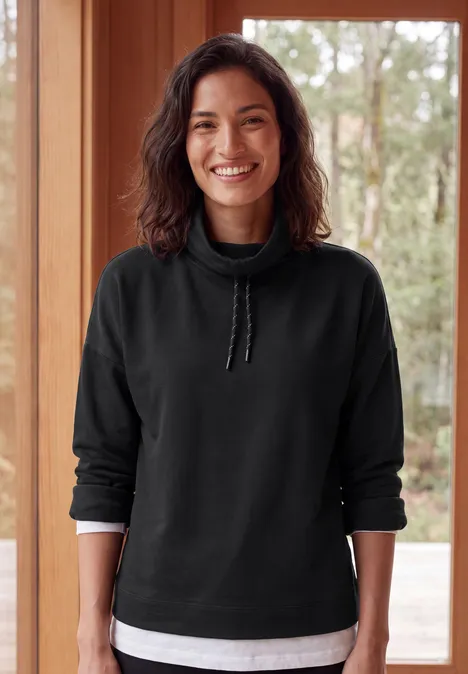 Shirt met lange mouwen, col en trekkoord Black Een lachende vrouw in een zwart sweatshirt en wit T-shirt staat voor een houten raam.
