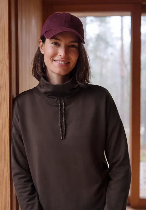 Langarm Shirt mit Rollkragen und Tunnelzug truffle brown Frau mit roter Kappe und dunklem Pullover lächelt vor hellem Fenster in Holzinterieur.