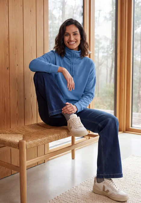 Langarm Shirt mit Rollkragen und Tunnelzug north blue Frau in blauem Rollkragenpullover und Jeans sitzt lächelnd auf einer Bank vor großen Fenstern.