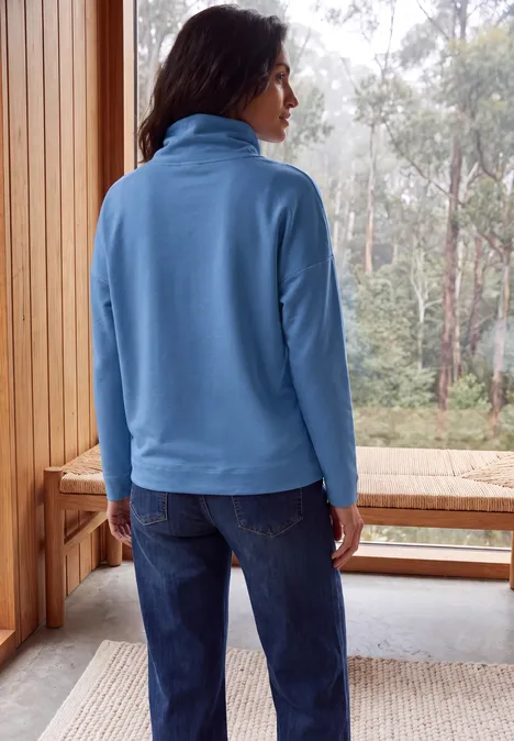 Langarm Shirt mit Rollkragen und Tunnelzug north blue Frau in blauer Fleecejacke und Jeans steht auf Holzveranda, schaut in nebligen Wald.